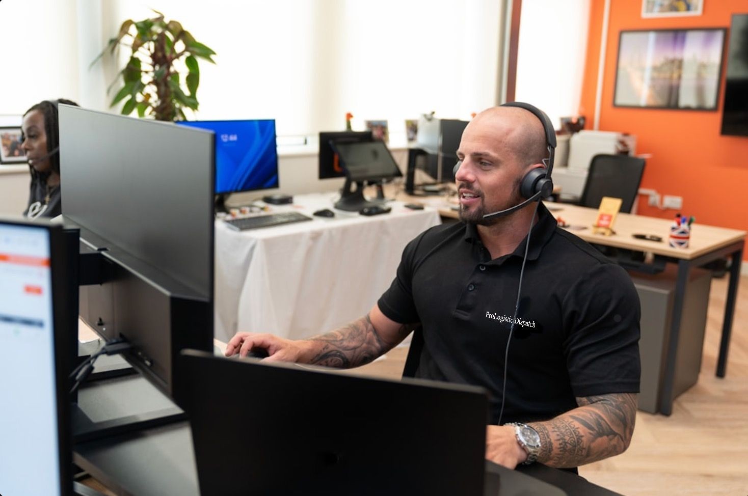 Man with a headset working at a dual-monitor computer desk in an office environment.