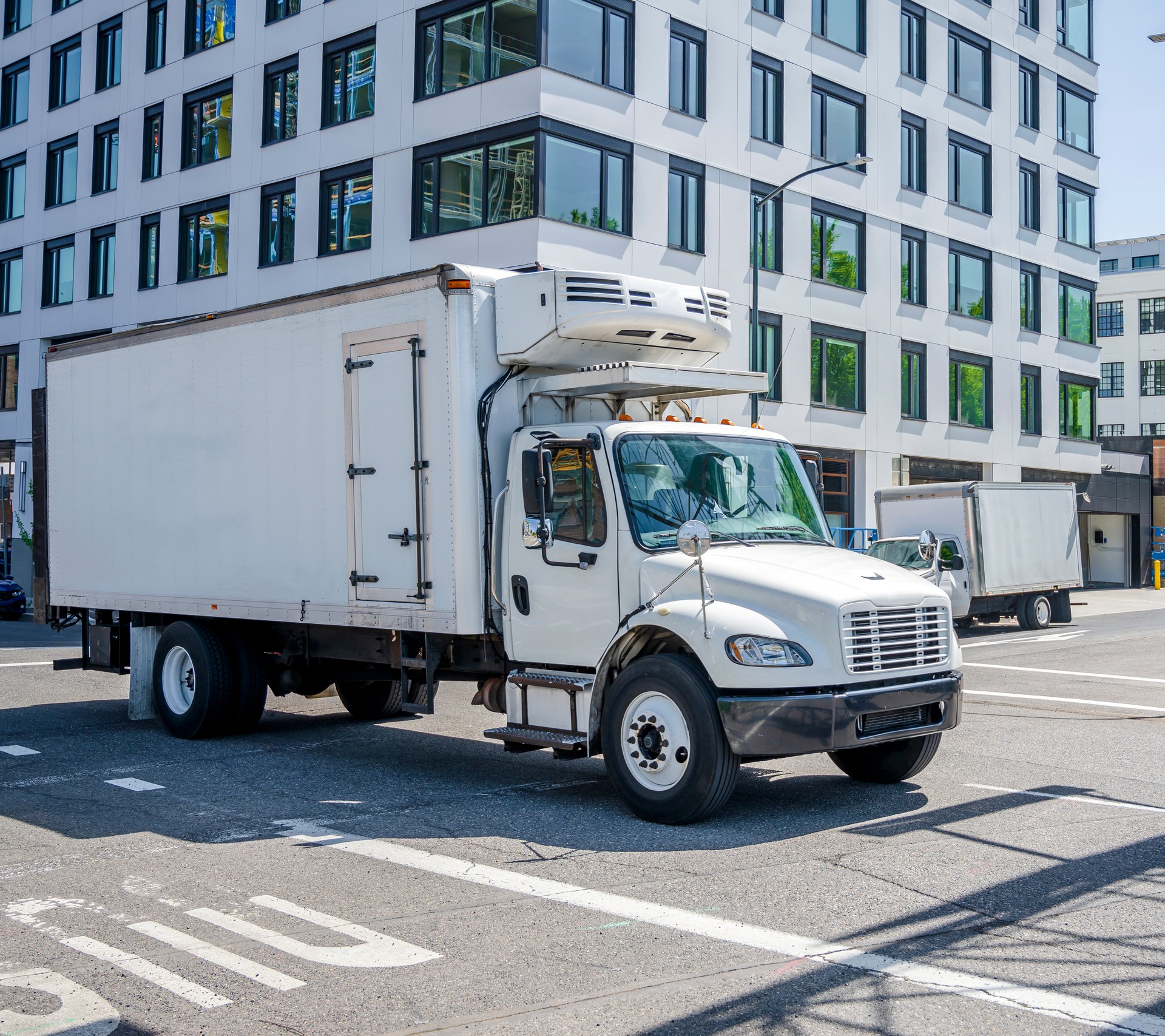 Small compact rig semi truck with refrigerator box trailer transporting goods on the street of urban city with multilevel buildings